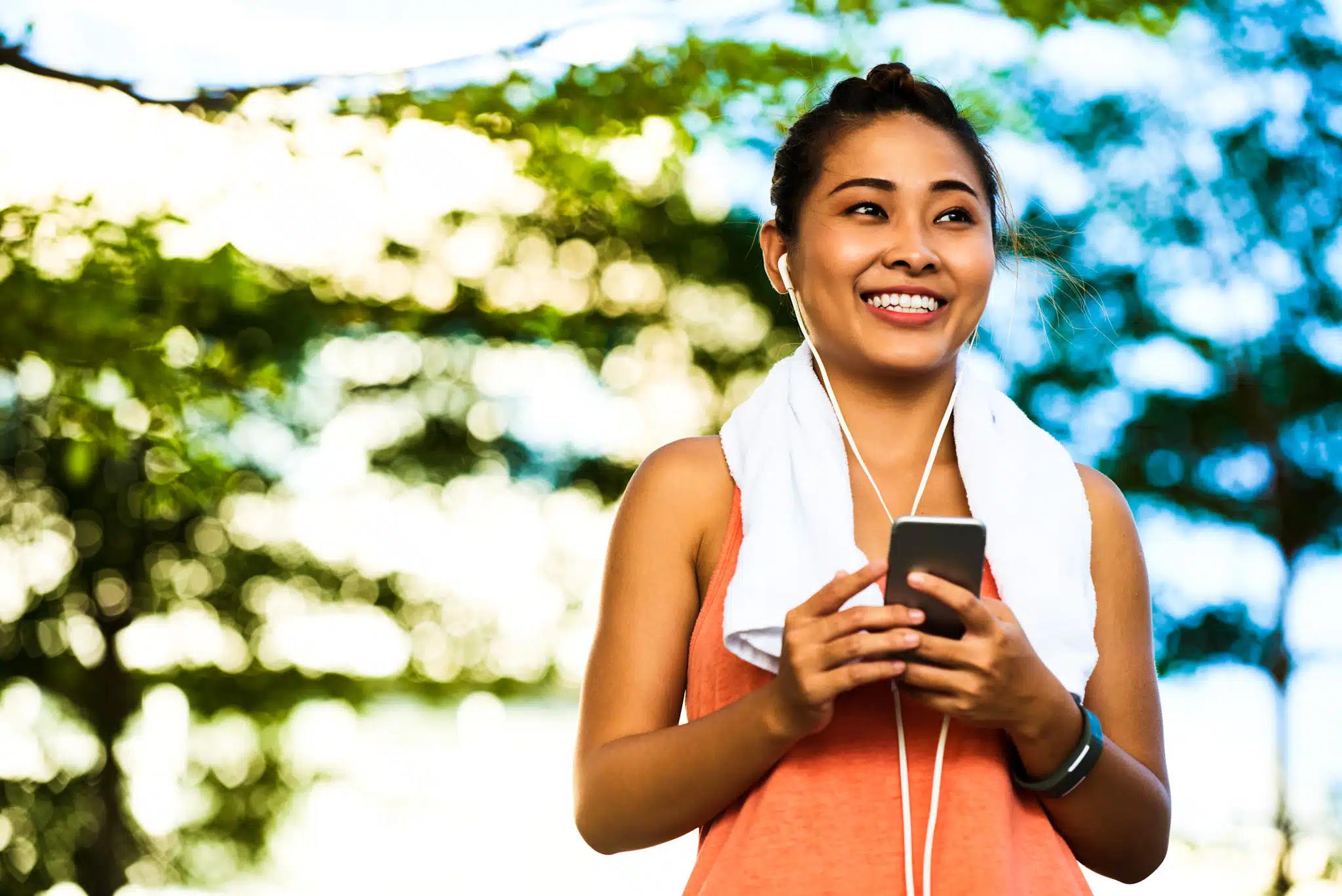A woman stands outdoors with earbuds in, smiling and holding a smartphone. A towel hangs around her neck. The background shows blurry trees and sunlight.