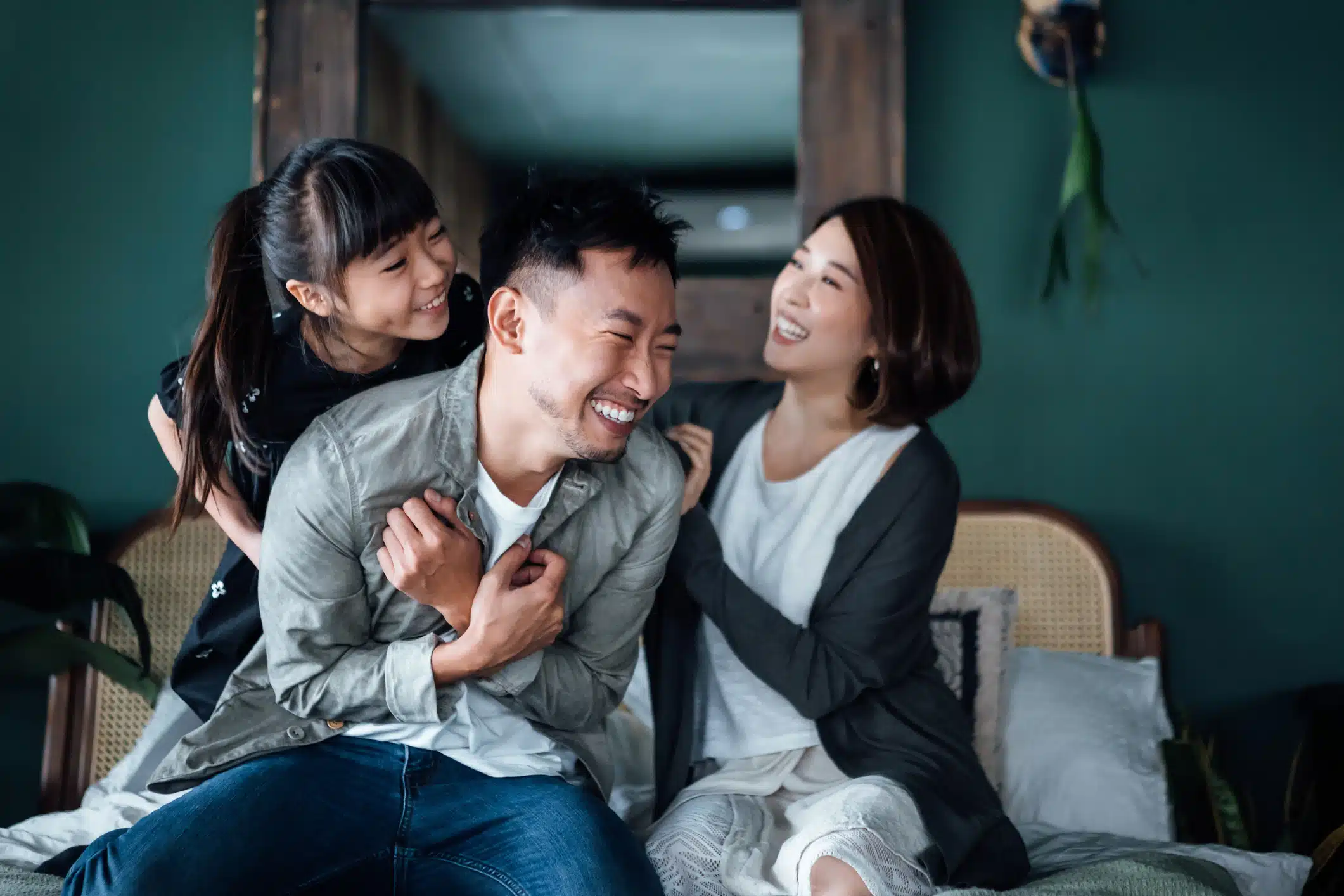 A cheerful man sits on a bed with his daughter and wife, all smiling and laughing in a cozy room with green walls.