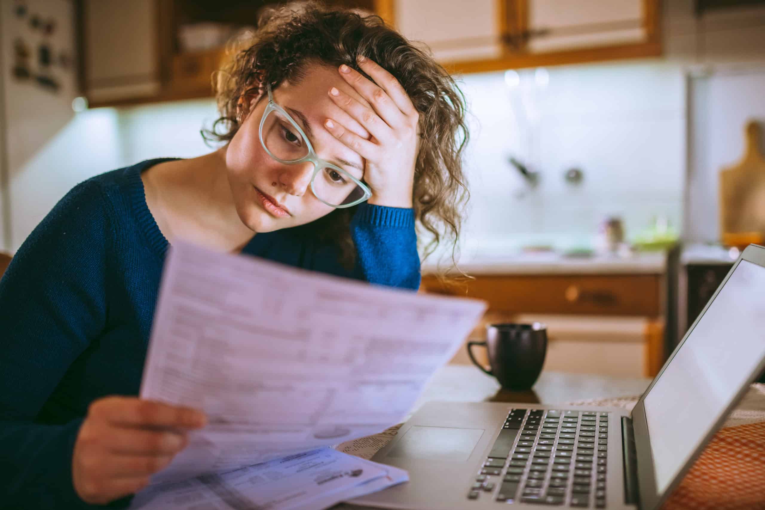 A person with curly hair and glasses looks at documents with a worried expression, sitting at a table with a laptop and coffee mug in a kitchen.