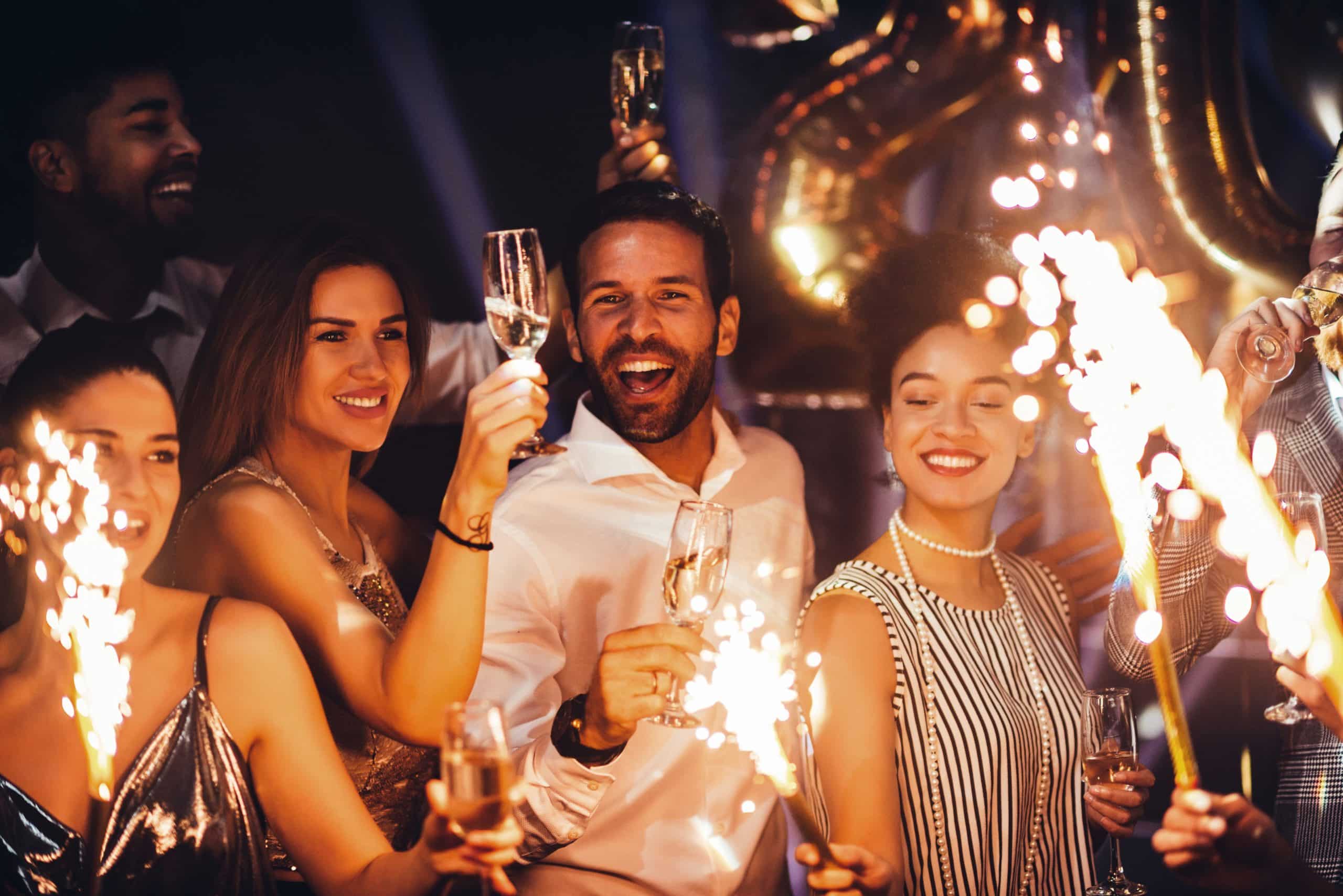 A group of people celebrating with champagne glasses and sparklers, surrounded by golden balloons.