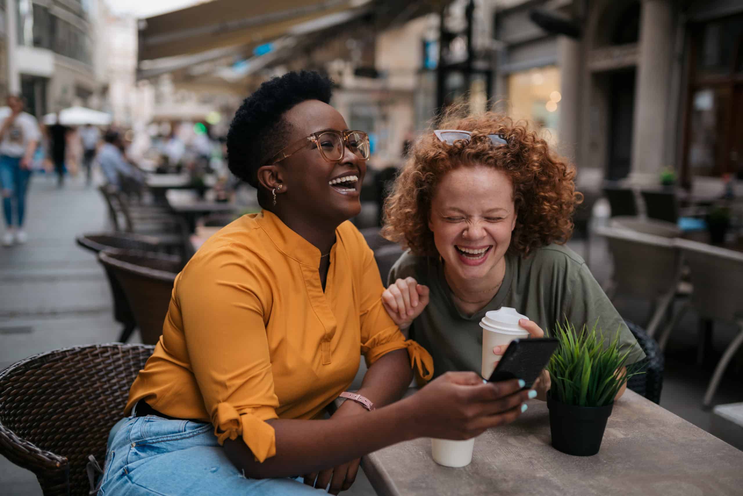 Two people sitting at an outdoor café, laughing together while looking at a phone. One person is holding a cup of coffee, and there are tables and other people in the background.