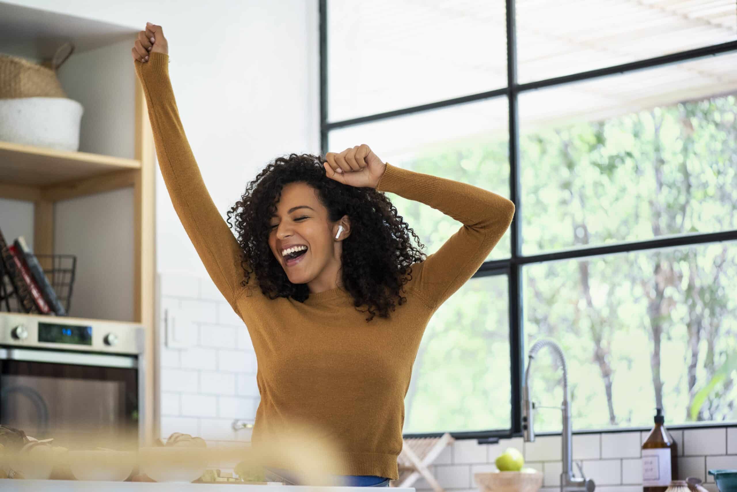 A woman stands in a kitchen with her arms raised, smiling. She has curly hair, is wearing a brown sweater, and has earbuds in her ears. Large windows and kitchen items are visible in the background.