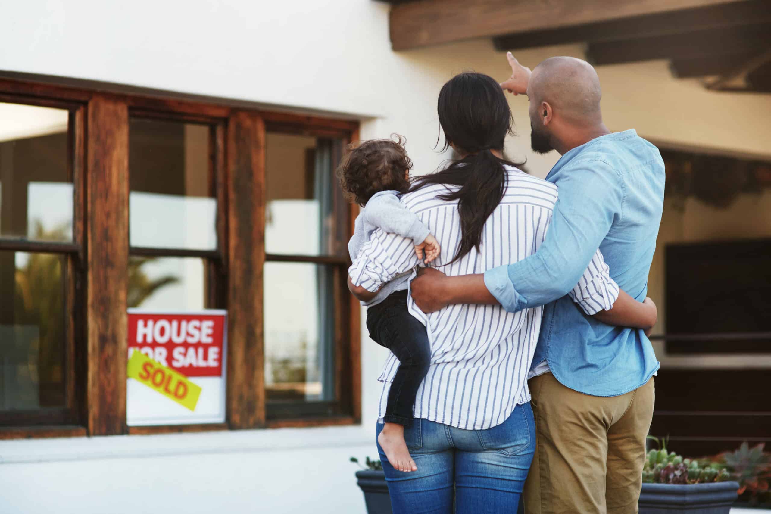 A family of three stands in front of a house with a "House for Sale" sign marked as "Sold" visible in the window.