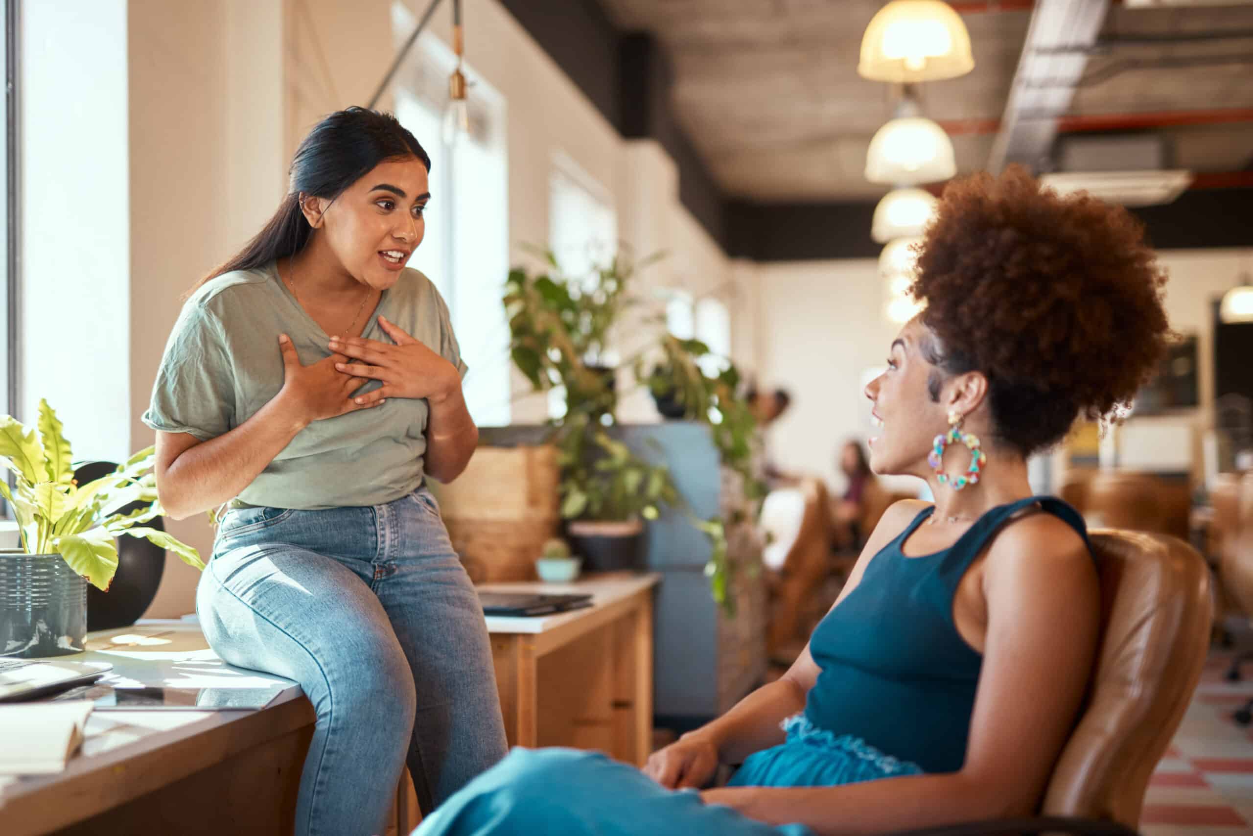 Two women are engaged in a conversation in a brightly lit room with plants and modern decor. One woman is seated on a desk, speaking earnestly, while the other listens attentively from a chair.