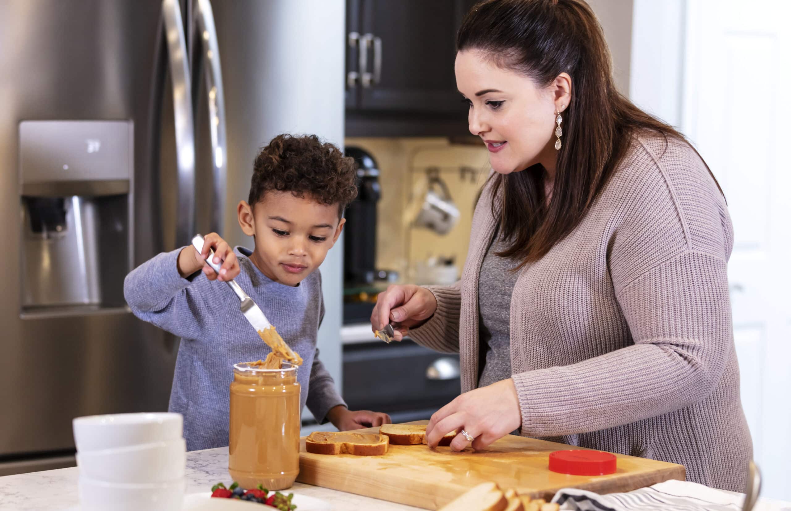 A woman and a child are in a kitchen spreading peanut butter on bread slices. The child is holding a butter knife, and the woman is assisting. An open jar of peanut butter is on the counter.