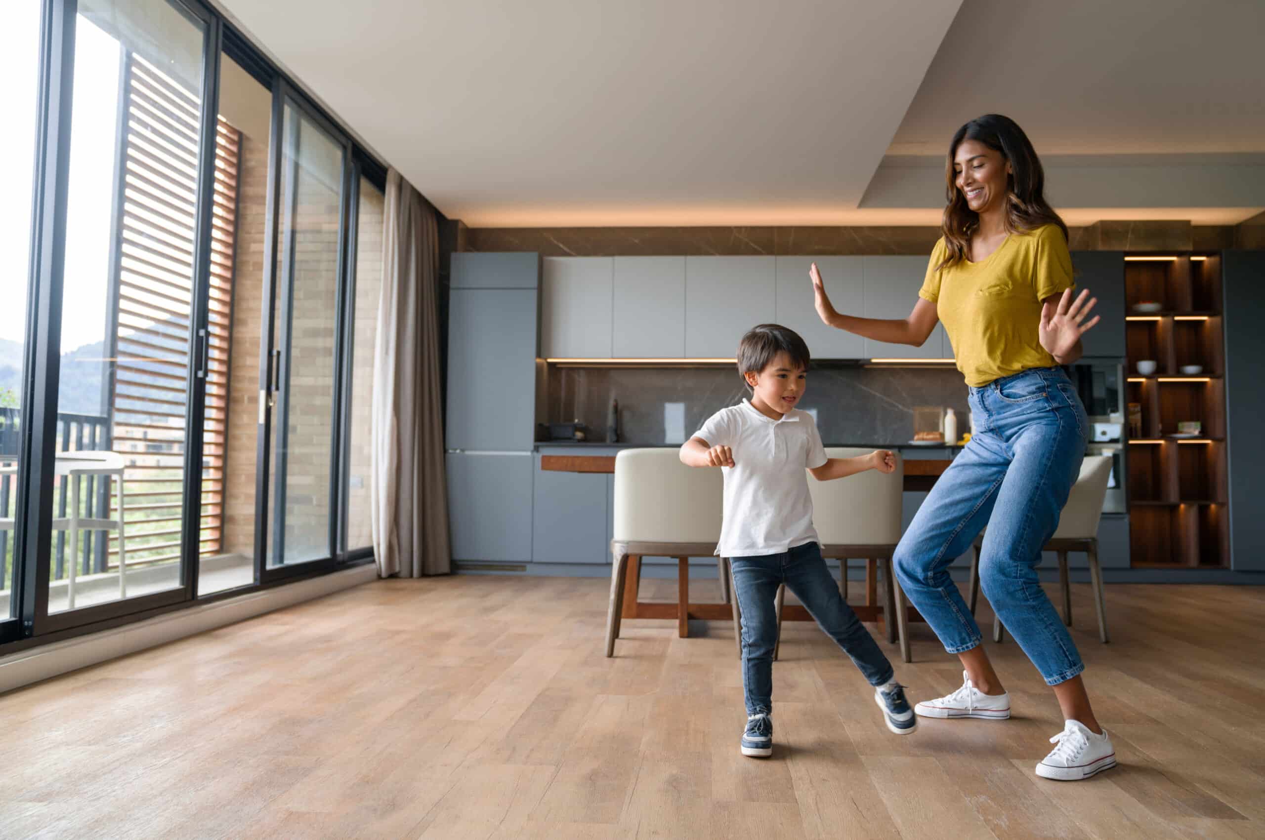 A woman and a young boy are dancing together in a spacious, modern kitchen with large windows and wooden flooring.