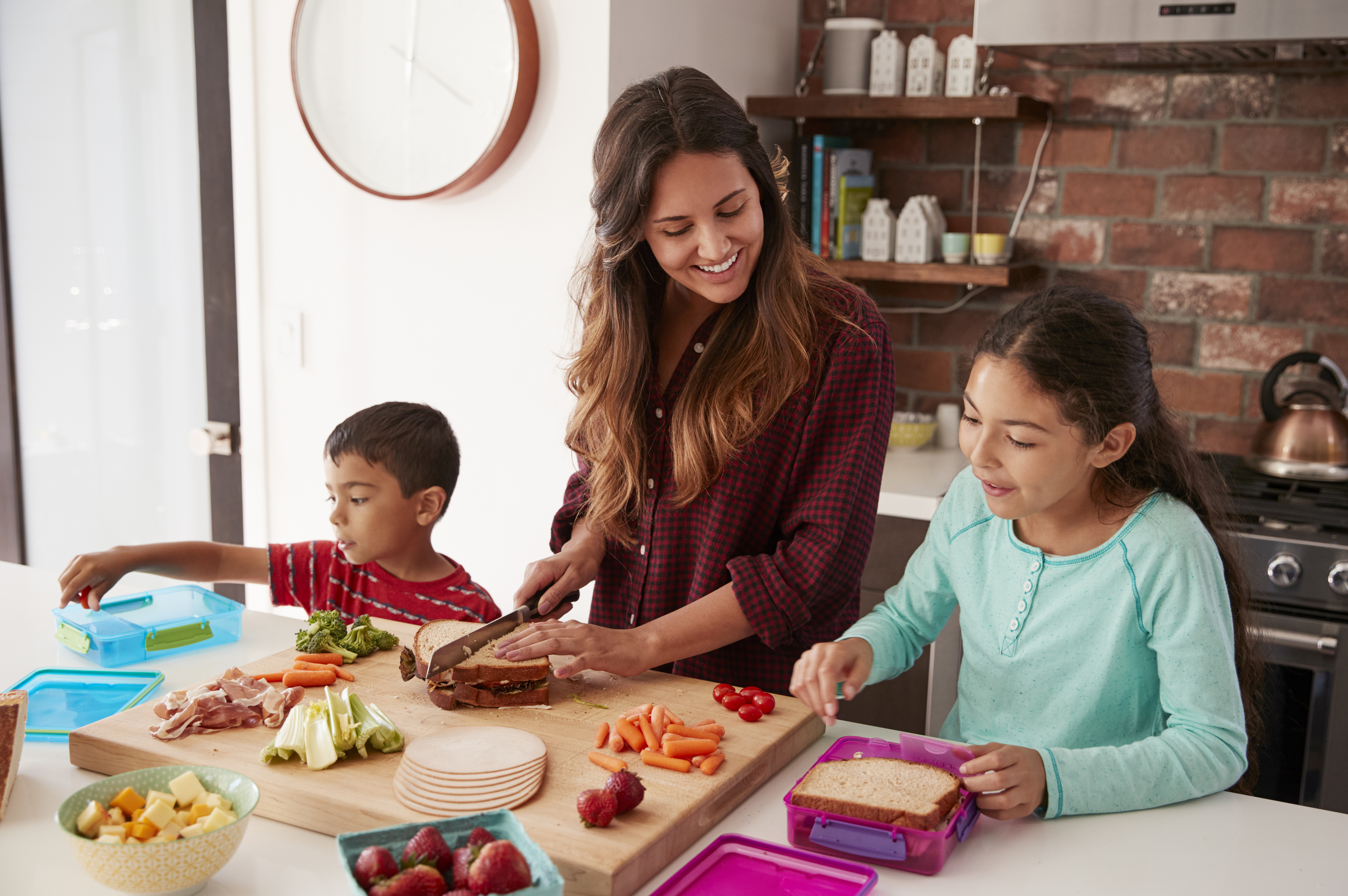 A woman prepares food with two children at a kitchen counter. She slices vegetables while the children pack lunch boxes with sandwiches and snacks.