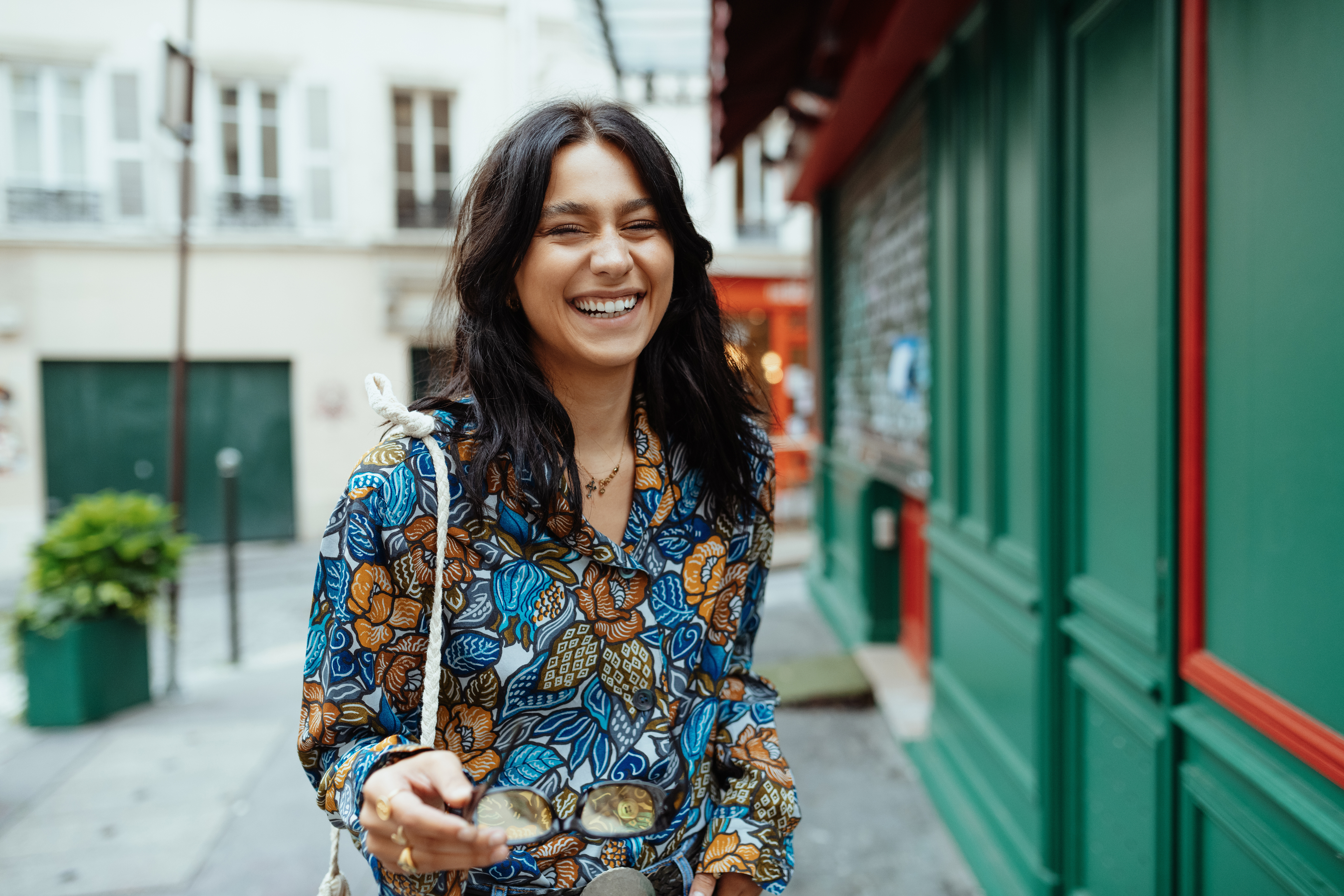 A person is smiling while standing on a city street, holding sunglasses and wearing a colorful patterned shirt.