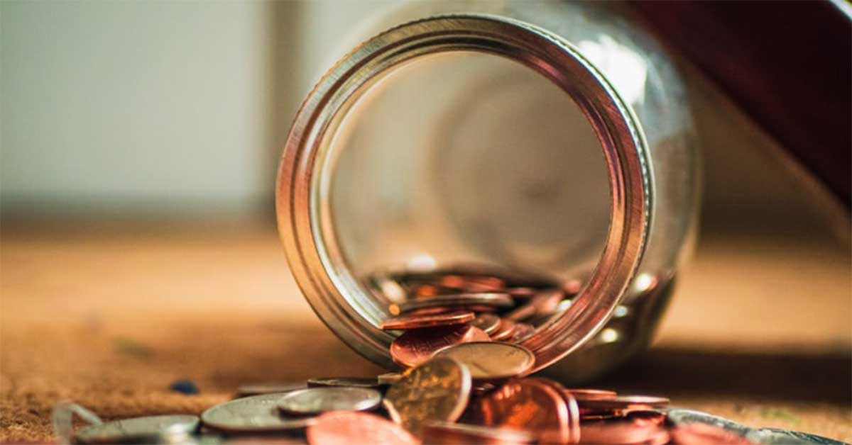 A clear jar tipped over on its side with assorted coins spilling out onto a surface.