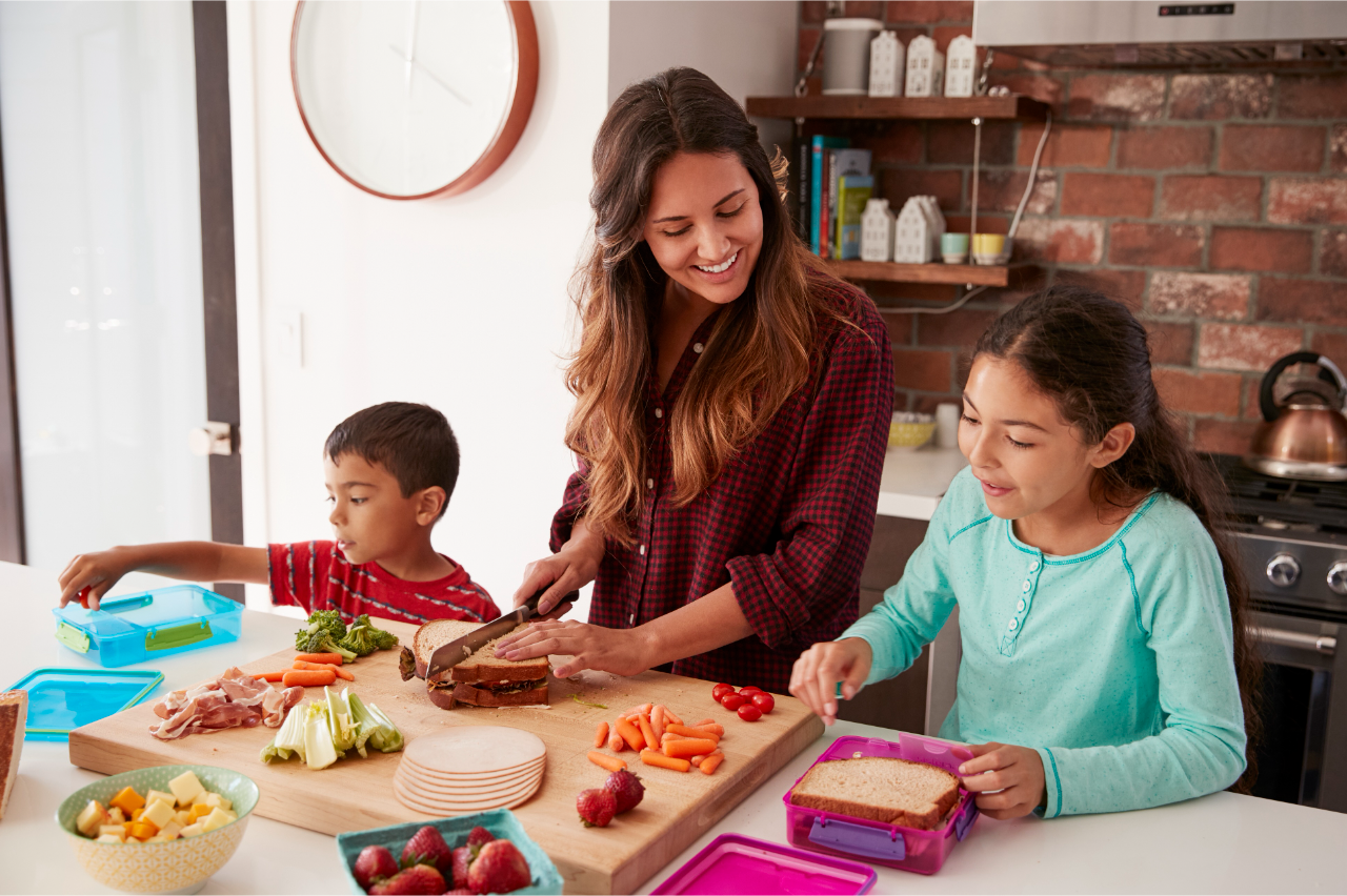 A woman is preparing sandwiches at a kitchen counter with a boy and a girl. The boy is packing food into a lunchbox, and the girl is spreading something on bread. Various ingredients are on the counter.