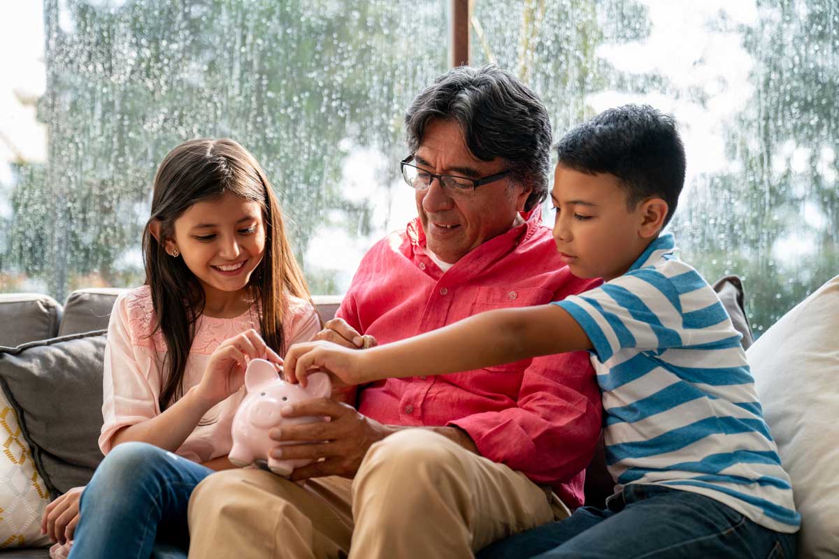 A man sits with a boy and a girl on a sofa, holding a pink piggy bank. They are indoors near a window with raindrops.