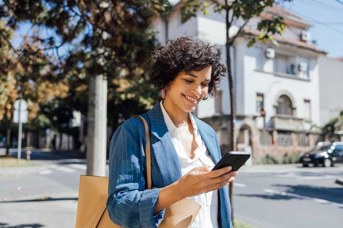 A smiling person in a blue blazer looks at their phone while standing on a tree-lined street, with buildings in the background.