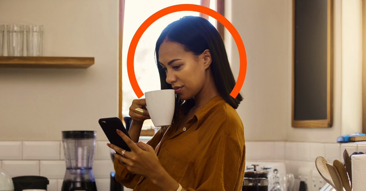 A woman stands in a kitchen, holding a white mug and looking at her phone. An orange semi-circle graphic is placed behind her head.